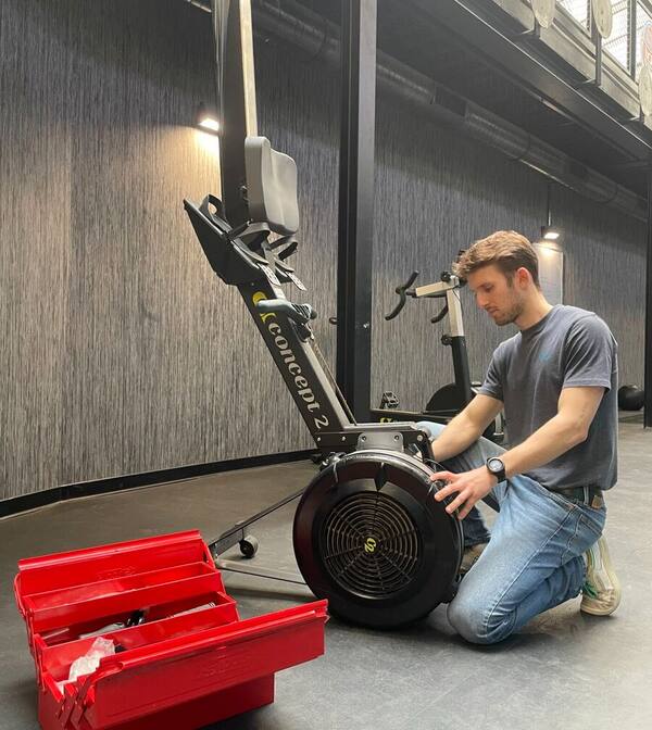 Technicien Ergo&Lift Maintenance réparant un rameur RowErg Concept 2 avec boîte à outils dans une salle de sport en Île-de-France