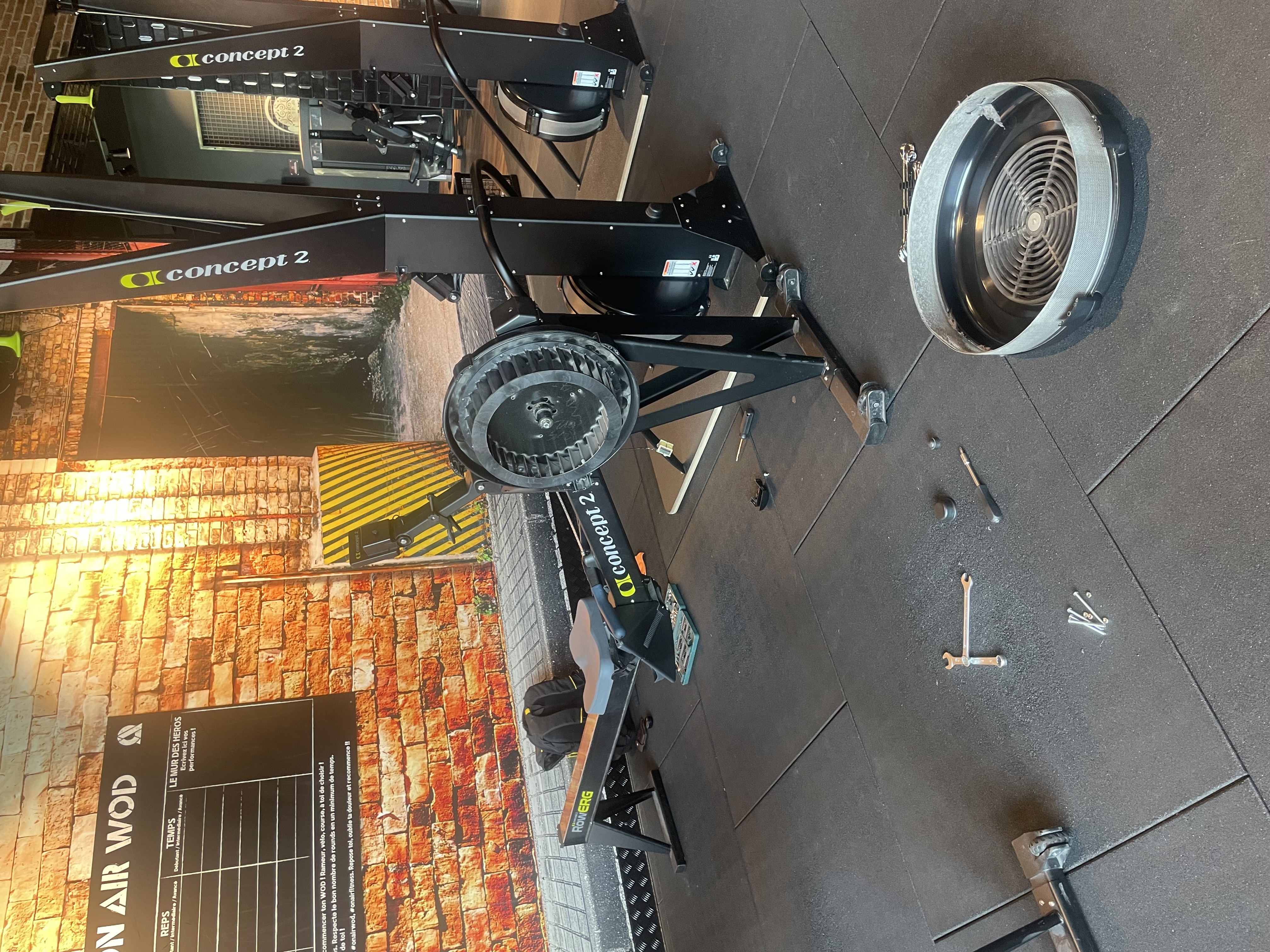 Rameur Concept 2 RowERG démonté en intervention — flywheel et carter séparés, outils au sol dans une salle de fitness